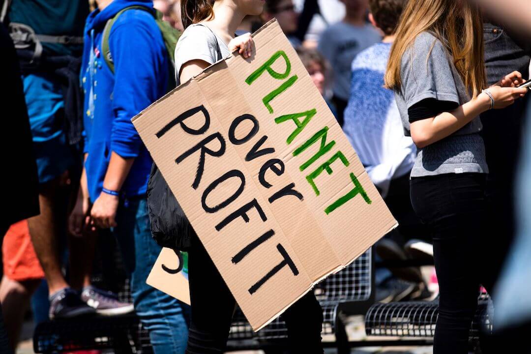 A young woman holds a handmade cardboard sign reading "Planet Over Profit".